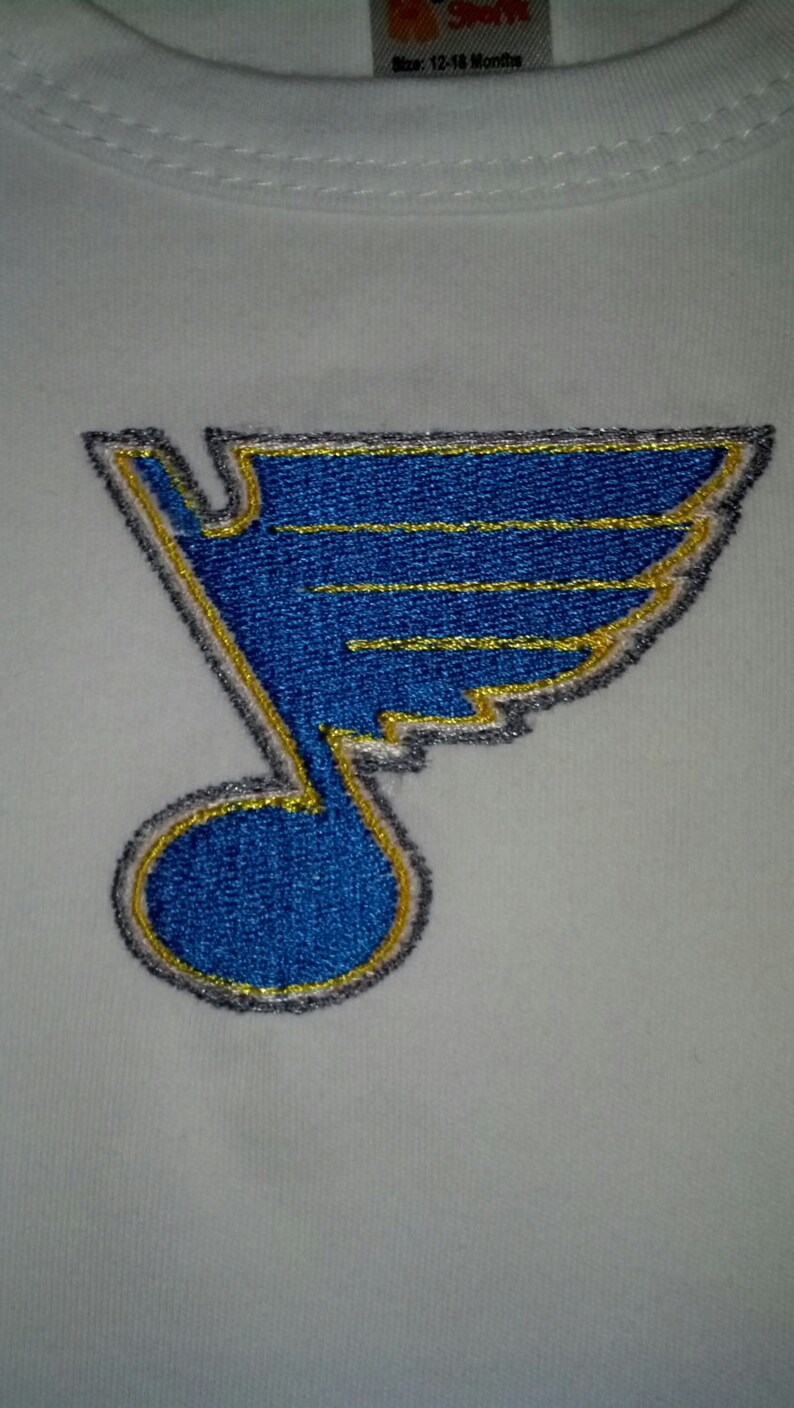 Pu&ograve; includere: Body bianco per neonato con un logo ricamato dei St. Louis Blues in blu e oro. Il logo &egrave; una nota musicale stilizzata con un'ala.