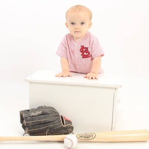 May include: A young child wearing a red and white baseball jersey with the letter "L" on the front, sits on a white wooden box. A baseball bat and a baseball glove are in front of the box.
