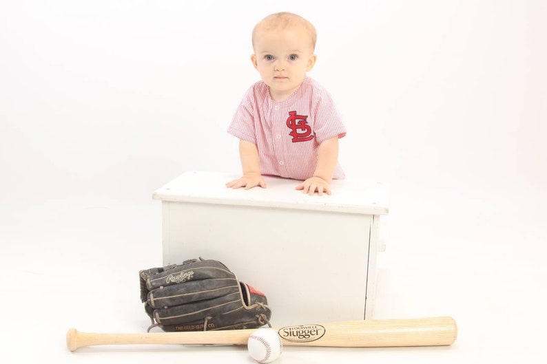 May include: A young child wearing a red and white baseball jersey with the letter "L" on the front, sits on a white wooden box. A baseball bat and a baseball glove are in front of the box.
