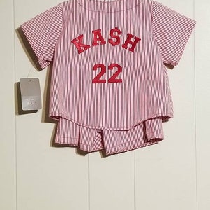 May include: Red and white striped baseball jersey with the name "KASH" and the number "22" embroidered on the back.