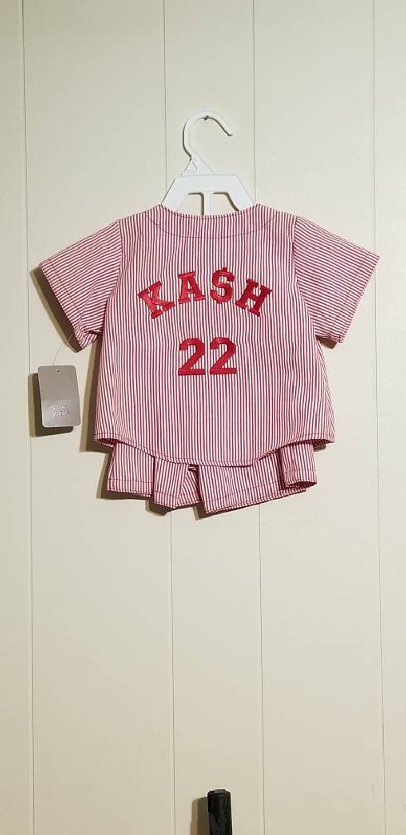 May include: Red and white striped baseball jersey with the name "KASH" and the number "22" embroidered on the back.