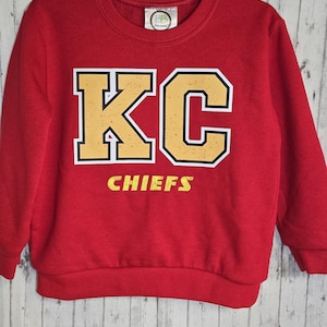 Può includere: Una felpa rossa con le lettere "KC" in oro e bianco, con la parola "CHIEFS" in oro sotto. La felpa ha un girocollo e maniche lunghe. Una piccola etichetta è visibile sul retro del collo.