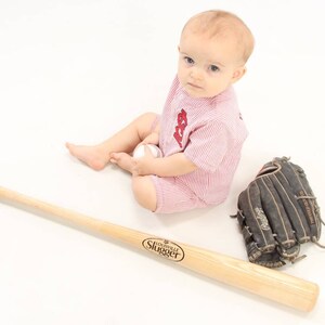 May include: A baby wearing a red and white striped baseball onesie is sitting on a white surface. The baby is holding a white baseball and looking at the camera. A wooden baseball bat and a black leather baseball glove are on the surface.