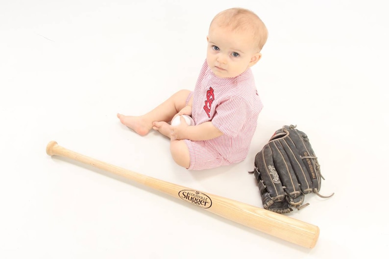 May include: A baby wearing a red and white striped baseball onesie is sitting on a white surface. The baby is holding a white baseball and looking at the camera. A wooden baseball bat and a black leather baseball glove are on the surface.