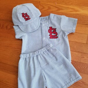 May include: A blue and white striped baseball uniform set for a baby. The set includes a short-sleeved shirt with a red 'STL' logo, matching shorts, and a blue and white striped baseball cap with a red 'STL' logo.