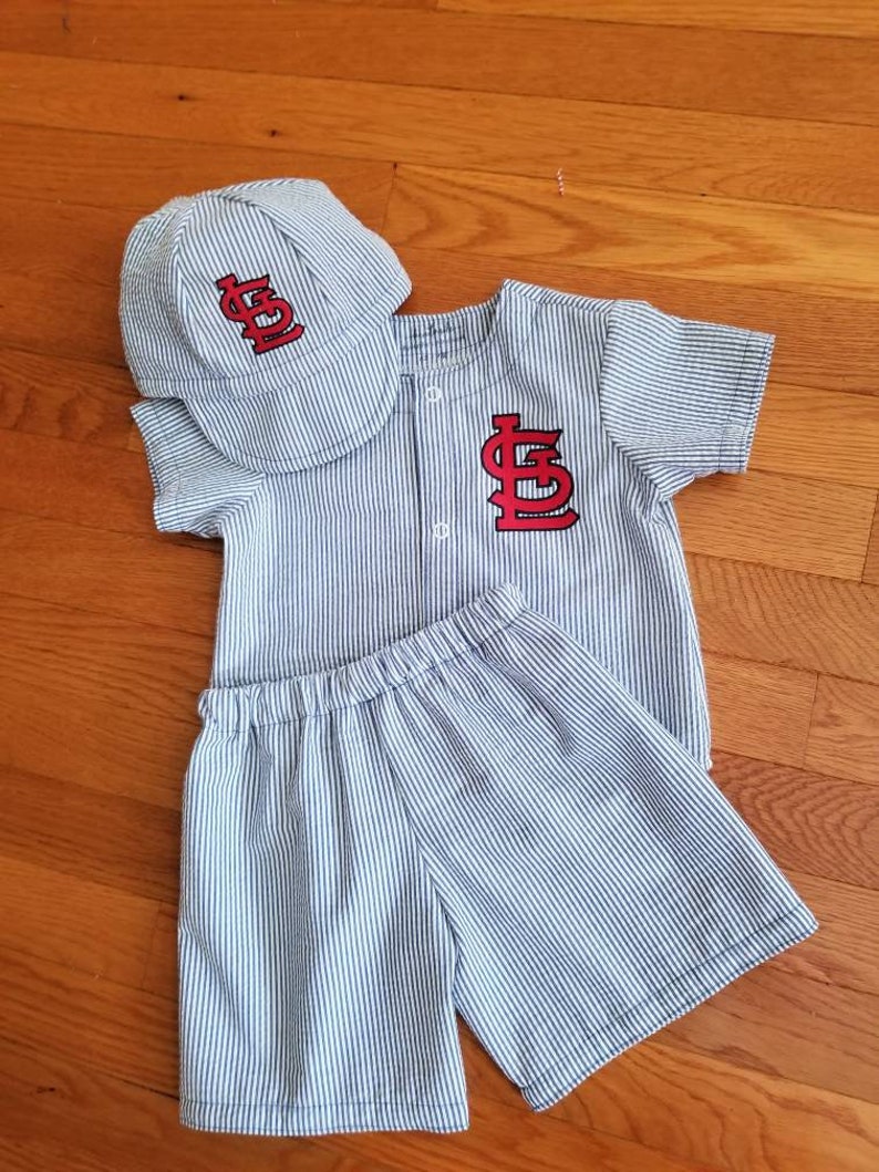 May include: A blue and white striped baseball uniform set for a baby. The set includes a short-sleeved shirt with a red 'STL' logo, matching shorts, and a blue and white striped baseball cap with a red 'STL' logo.