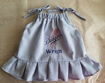 May include: A blue and white striped sleeveless dress with a ruffled hem. The dress has an embroidered baseball logo with the text "Dodgers" and the name "Wren" underneath.