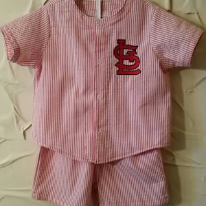 May include: A red and white striped short-sleeved shirt and matching shorts set for a baby or toddler. The shirt has a red embroidered baseball logo with the letters "LS" on the front.