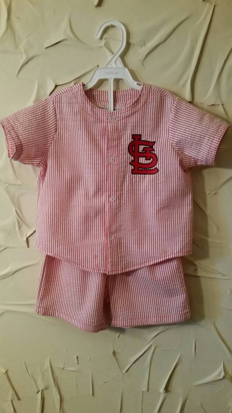 May include: A red and white striped short-sleeved shirt and matching shorts set for a baby or toddler. The shirt has a red embroidered baseball logo with the letters "LS" on the front.