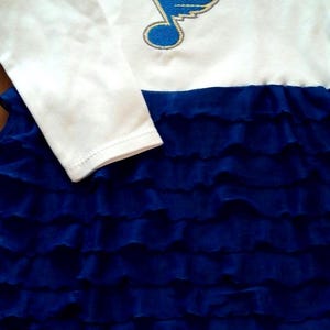 May include: White long-sleeved baby onesie with a blue and gold embroidered St. Louis Blues logo. The onesie has a blue ruffled skirt attached.