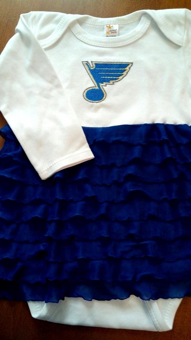 May include: White long-sleeved baby onesie with a blue and gold embroidered St. Louis Blues logo. The onesie has a blue ruffled skirt attached.
