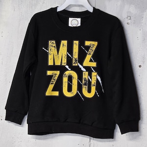 Puede incluir: Sudadera negra de cuello redondo con el texto "MIZ ZOU" en amarillo, con letras desgastadas. Rayas blancas acentúan el texto. La sudadera está colgada en una percha blanca sobre un fondo gris texturizado.