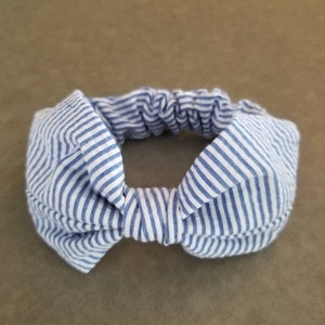 May include: A blue and white striped headband with a bow in the center. The headband is made of a soft, stretchy fabric.