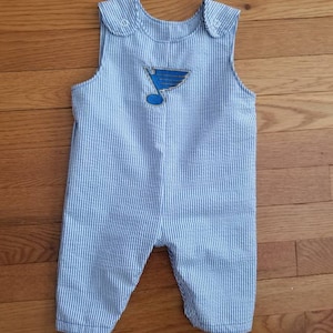 Könnte beinhalten: Blaue und weiße gestreifte Baby-Latzhosen mit einem gestickten St. Louis Blues-Logo in Blau und Gold auf der Vorderseite.