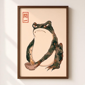 May include: A framed wall art print featuring a grumpy, dark green mottled frog with a cream-colored belly. The frog holds a small brown bowl and a pestle. In the upper left corner, there is a red rectangular stamp containing the Japanese text 問一.