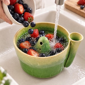 May include: A green ceramic berry colander bowl with a central drainage post and a side spout. The bowl features a gradient glaze from light green at the rim to a darker green base, currently filled with water, fresh strawberries, and blueberries.