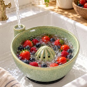 Può includere: Una ciotola scolapasta per frutti di bosco in ceramica verde chiaro con finitura maculata e una cupola di drenaggio centrale. La ciotola è riempita d'acqua e fragole, lamponi e mirtilli freschi, posizionata sotto il getto di un rubinetto.