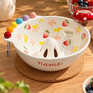 May include: A white ceramic fruit bowl featuring a central juicer cone, painted with red strawberries and yellow and pink flowers. The rim includes four colorful spheres, a red mushroom-shaped holder, and the word name in red script.