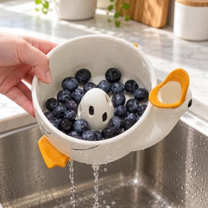 May include: A white ceramic colander shaped like a duck with orange accents, featuring a central drainage cone and side spout. The bowl is filled with fresh blueberries, and water is seen draining through the bottom holes into a sink.
