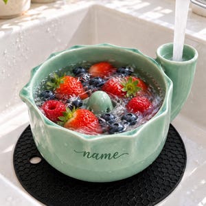 May include: A light green ceramic berry colander bowl with a scalloped rim and a side spout for water drainage. The bowl features the word name in cursive script and is filled with water, fresh strawberries, and blueberries under a faucet.