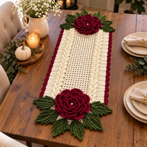 May include: A rectangular crochet table runner featuring a cream-colored lattice center bordered by braided cream yarn. Each end is decorated with a large, deep red crocheted rose with a central pearl accent, surrounded by dark green crocheted leaves.