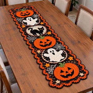 Peut inclure: Un chemin de table en crochet rectangulaire présentant un motif d'Halloween répétitif composé de citrouilles orange, de fantômes blancs et d'un chat noir. Le design inclut des toiles d'araignées blanches et de petites étoiles jaunes, bordé d'un feston noir.