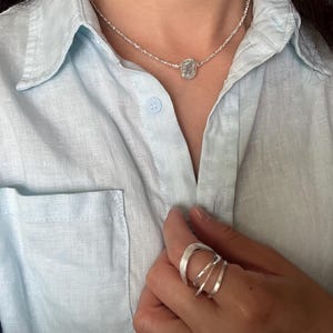 Könnte beinhalten: Eine silberne Halskette mit einer feinen Kette und einem kleinen, strukturierten, ovalen Anhänger, kombiniert mit einem silbernen Ring aus drei glatten, überlappenden und leicht geschwungenen Bändern am Finger.