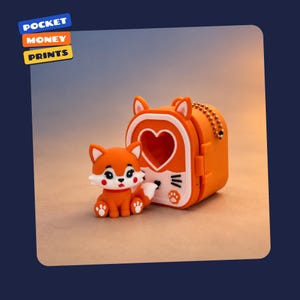 May include: An orange fox figurine with white accents and a matching orange box featuring a heart-shaped cutout, cat-like whiskers, and a paw print. The box includes a gold-coloured ball chain. The text POCKET MONEY PRINTS appears in the corner.