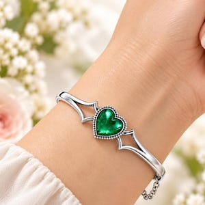 May include: A silver-toned cuff bracelet featuring a central heart-shaped green stone with a beaded border. The metal band has a pointed, angular design on both sides of the heart and includes a small chain link section for closure.