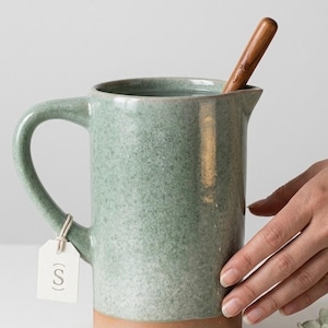 May include: A sage green ceramic pitcher with a speckled glaze and a natural clay base. A wooden stirring stick rests inside the pitcher. A small white rectangular tag with the letter S printed in a circle is attached to the handle with string.
