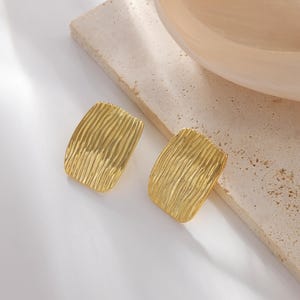 May include: A pair of gold-toned stud earrings featuring a rectangular shape with rounded corners and a textured, wavy, rippled surface pattern. The earrings have a polished metallic finish and a flat, solid construction on the front-facing side.
