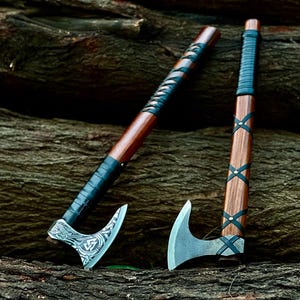 May include: Two axes with wooden handles and metal heads. The left axe features a silver-toned head with intricate etched patterns and a black leather-wrapped handle. The right axe has a plain metal head and a handle with black leather cross-lacing.