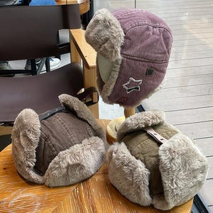 May include: Three trapper hats with faux fur lining and ear flaps. One hat is mauve with a star patch, one is dark brown, and one is olive green. Each hat features a top strap with a metal snap button closure and textured fabric exterior.