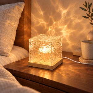 Peut inclure: Une lampe de table carrée dotée d'un abat-jour en cube de verre transparent texturé qui émet une lumière dorée et chaude. Le verre présente un motif de surface irrégulier et ondulé. La lampe repose sur une base carrée en bois clair avec un cordon.