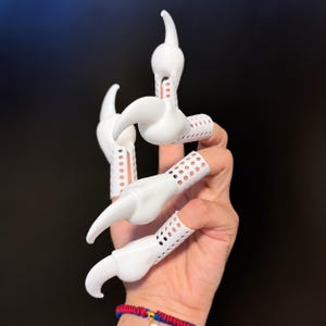 Puede incluir: Un conjunto de cuatro extensiones de dedo blancas con forma de garras afiladas y curvas. Cada pieza presenta una base perforada tipo malla que se ajusta al dedo, terminando en una punta de garra sólida, lisa y puntiaguda.