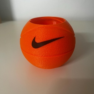 May include: A bright orange, spherical container with a textured surface resembling a basketball. The object features a black Nike swoosh logo embossed on the front and has an open top, displaying a hollow interior with visible 3D printed layers.