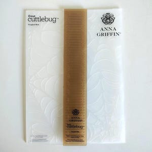 May include: A white rectangular Cricut Cuttlebug embossing folder with a Tangled Web pattern, featuring the Anna Griffin logo. A translucent brown plastic spacer plate is centred vertically over the folder, displaying Anna Griffin branding.