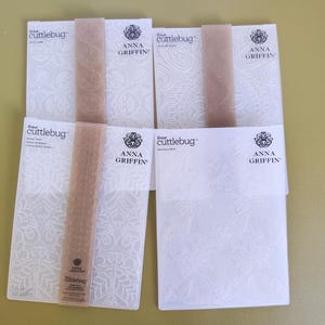 May include: Four rectangular white plastic embossing folders by Cricut Cuttlebug and Anna Griffin. Each folder features intricate embossed patterns, including floral and bell designs, with branded paper bands across the center of three.