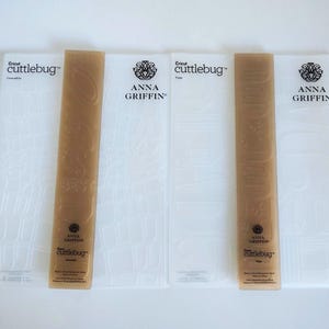 May include: Two sets of translucent plastic embossing folders by Cricut Cuttlebug and Anna Griffin. Each set includes a textured folder and a tan-colored strip with embossed text. The branding includes the Cricut Cuttlebug logo and the Anna Griffin name printed in black ink on the surfaces.