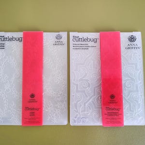 May include: Two rectangular Cricut Cuttlebug embossing folders by Anna Griffin. The left folder features a floral trellis pattern, while the right folder displays an intricate quatrefoil design. Each folder has a bright pink paper band.
