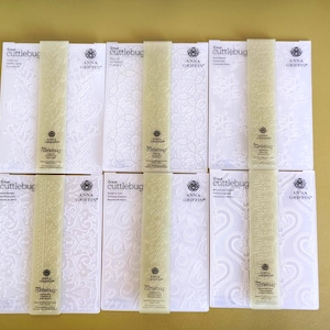May include: Six white rectangular embossing folders by Cricut Cuttlebug and Anna Griffin, each featuring distinct embossed floral and scroll patterns. The folders are arranged in two rows of three, with vertical branding strips centered on each.