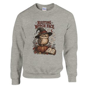 May include: A heather gray crewneck sweatshirt featuring a graphic of an owl wearing a witch hat, perched on a pumpkin. The text reads RESTING WITCH FACE above the owl, with an open book, autumn leaves, and stars surrounding the central design.