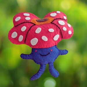May include: A felt plush toy shaped like a creature with a large, pink, mushroom-like head featuring white polka dots and an orange centre. The body is a rounded blue shape with two small arms and two legs, featuring a stitched smile and two red dots.