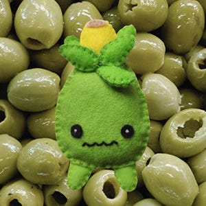 May include: A light green felt plush toy shaped like an olive with a yellow top, green leaves, two black circular eyes, and a stitched wavy mouth, positioned against a background of numerous pitted green olives.