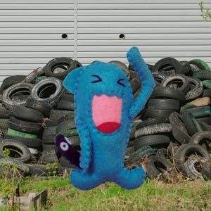 Peut inclure: Une peluche en feutrine bleue avec une bouche grande ouverte à l'intérieur rose, des yeux fermés représentés par des coutures sombres et une petite queue violet foncé avec une pointe circulaire blanche. Les bords présentent des points de feston.