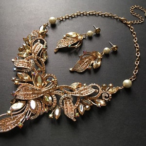 May include: A gold-toned necklace and earring set. The necklace features a floral design with clear and amber-colored crystals. The earrings have a leaf design with a pearl accent. The jewelry set is on a black background.