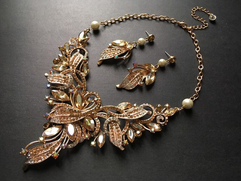 May include: A gold-toned necklace and earring set. The necklace features a floral design with clear and amber-colored crystals. The earrings have a leaf design with a pearl accent. The jewelry set is on a black background.