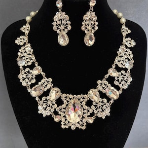 May include: A silver-toned necklace and earring set featuring clear, faceted crystals in teardrop and round shapes arranged in a floral-inspired filigree pattern. The necklace includes small white pearls along the chain and the text Weddingvalle.