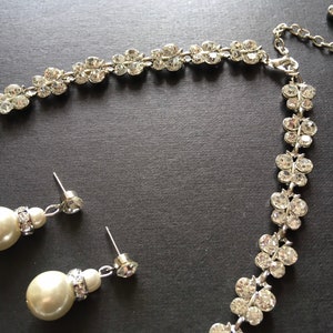 Puede incluir: Un conjunto de joyas plateadas con collar y pendientes. El collar y los pendientes est&aacute;n adornados con brillantes transparentes dispuestos en un patr&oacute;n floral. Los pendientes tienen perlas grandes, redondas y de color blanco roto.