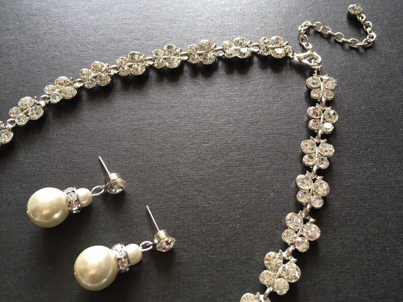 Puede incluir: Un conjunto de joyas plateadas con collar y pendientes. El collar y los pendientes est&aacute;n adornados con brillantes transparentes dispuestos en un patr&oacute;n floral. Los pendientes tienen perlas grandes, redondas y de color blanco roto.